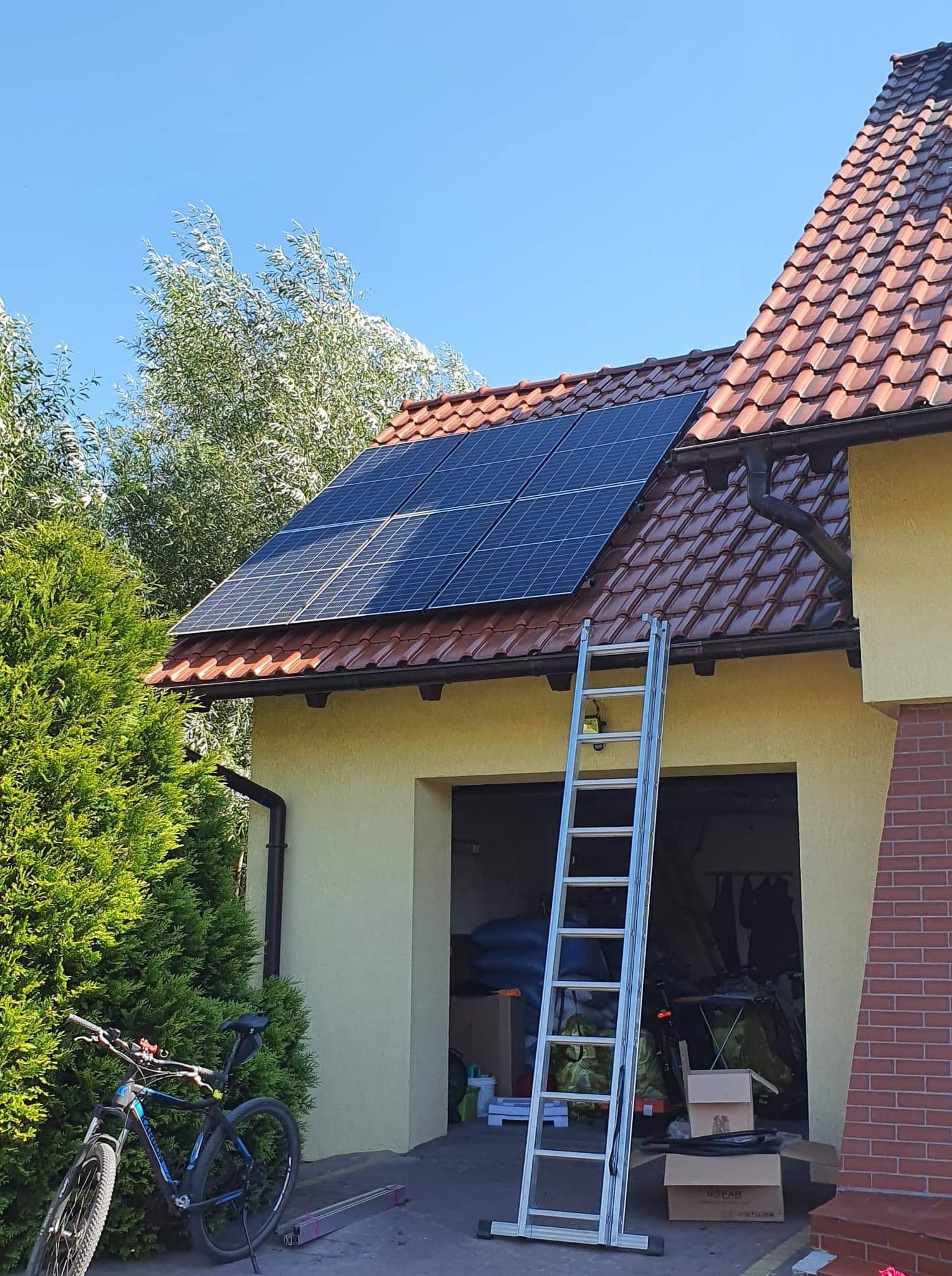 Panele fotowoltaiczne zamontowane na dachu domu z czerwonej dachówki, drabina oparta o rynnę, rower górski oparty o tuję, otwarty garaż z widocznym bałaganem w tle.