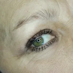 Zbliżenie na oko kobiety z widoczną delikatną kreską permanentną na górnej powiece, podkreślającą linię rzęs. Zielona tęczówka z wyraźnym odbiciem światła.