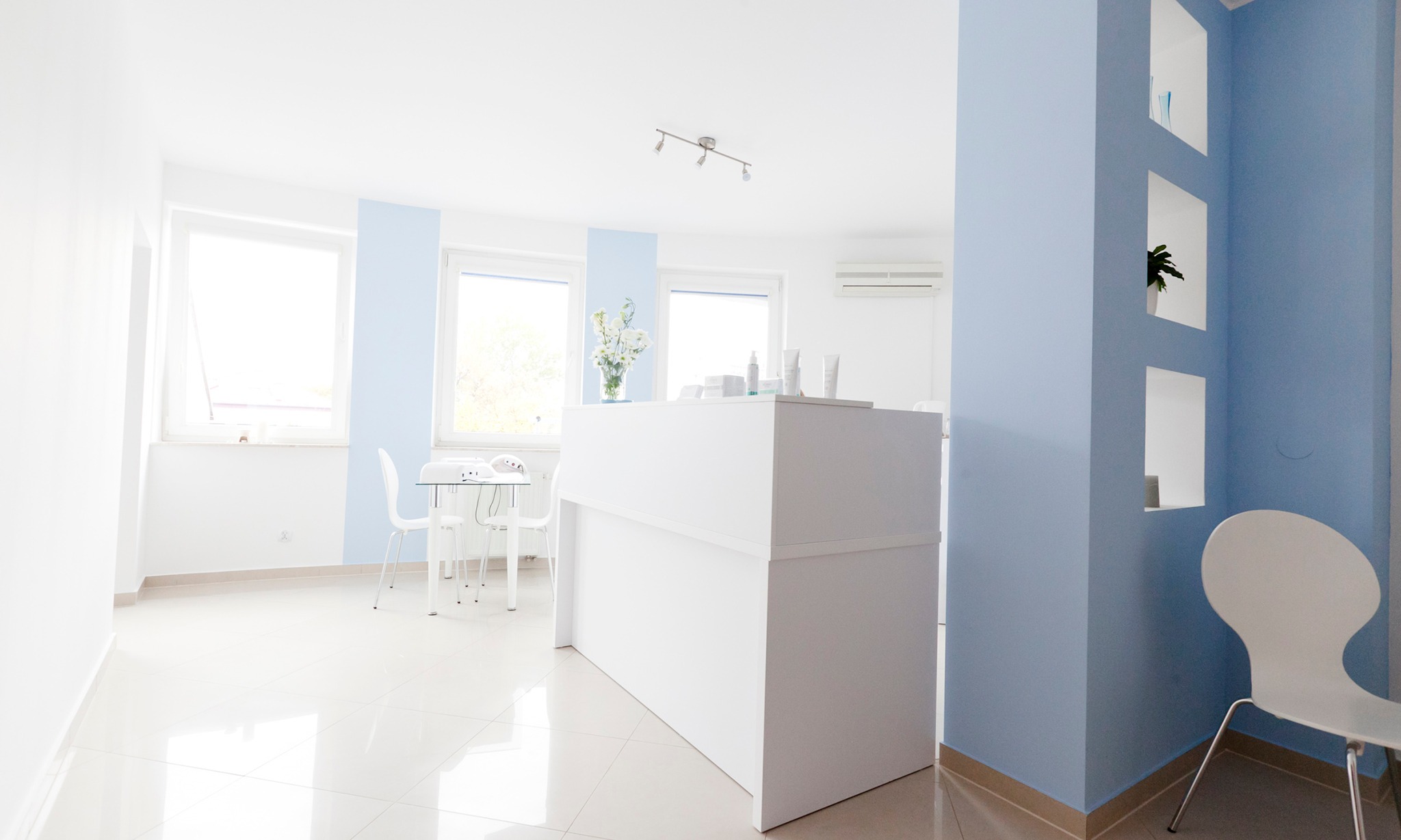 Jasne wnętrze salonu kosmetycznego z białą recepcją, stolikiem do manicure i błękitnymi akcentami na ścianach.