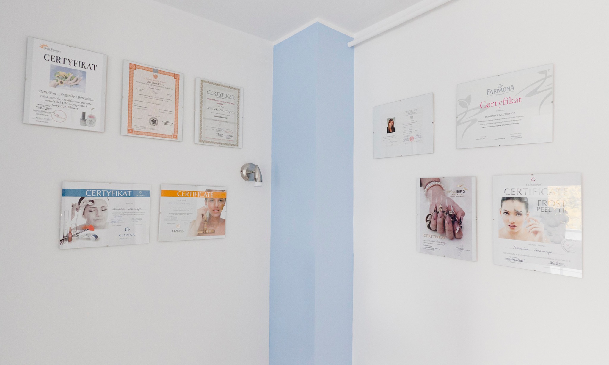 Ściana gabinetu kosmetycznego z powieszonymi certyfikatami ukończenia kursów i szkoleń z zakresu stylizacji paznokci, zabiegów na twarz i ciało, w ramkach z pleksi, oświetlona punktowym światłem.