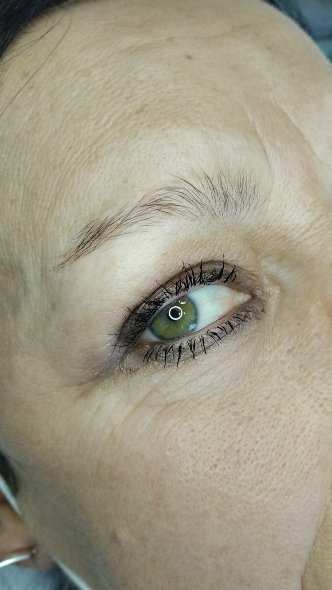 Zbliżenie na oko kobiety z widoczną delikatną kreską permanentną na górnej powiece, podkreślającą linię rzęs. Zielona tęczówka z wyraźnym odbiciem światła.