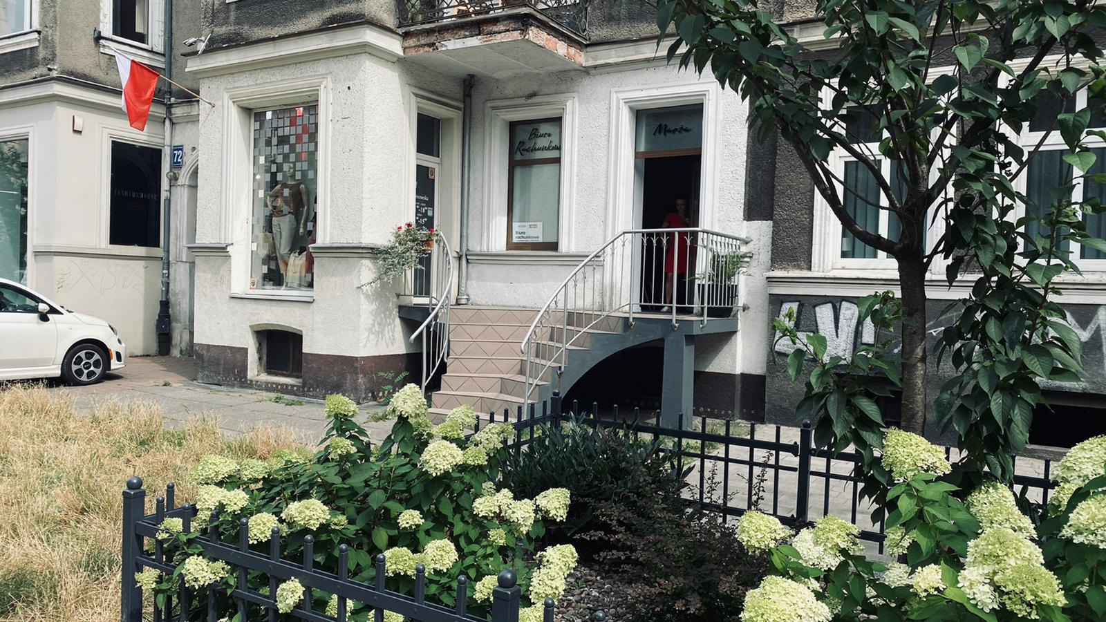 Fasada budynku biura rachunkowego z wejściem po schodach, widoczny szyld i flaga Polski