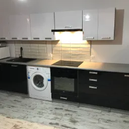 Nowoczesna kuchnia w Żninie z białymi szafkami górnymi i czarnymi dolnymi, z widoczną pralką w zabudowie, płytą indukcyjną i piekarnikiem.