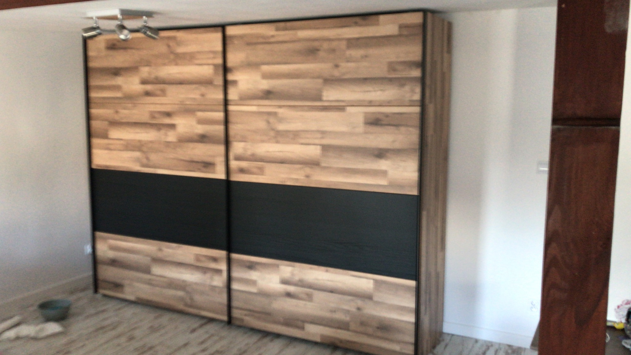 Dwudrzwiowa szafa przesuwna z drewnopodobnymi panelami i czarnym pasem pośrodku, oświetlona trzema reflektorami sufitowymi.