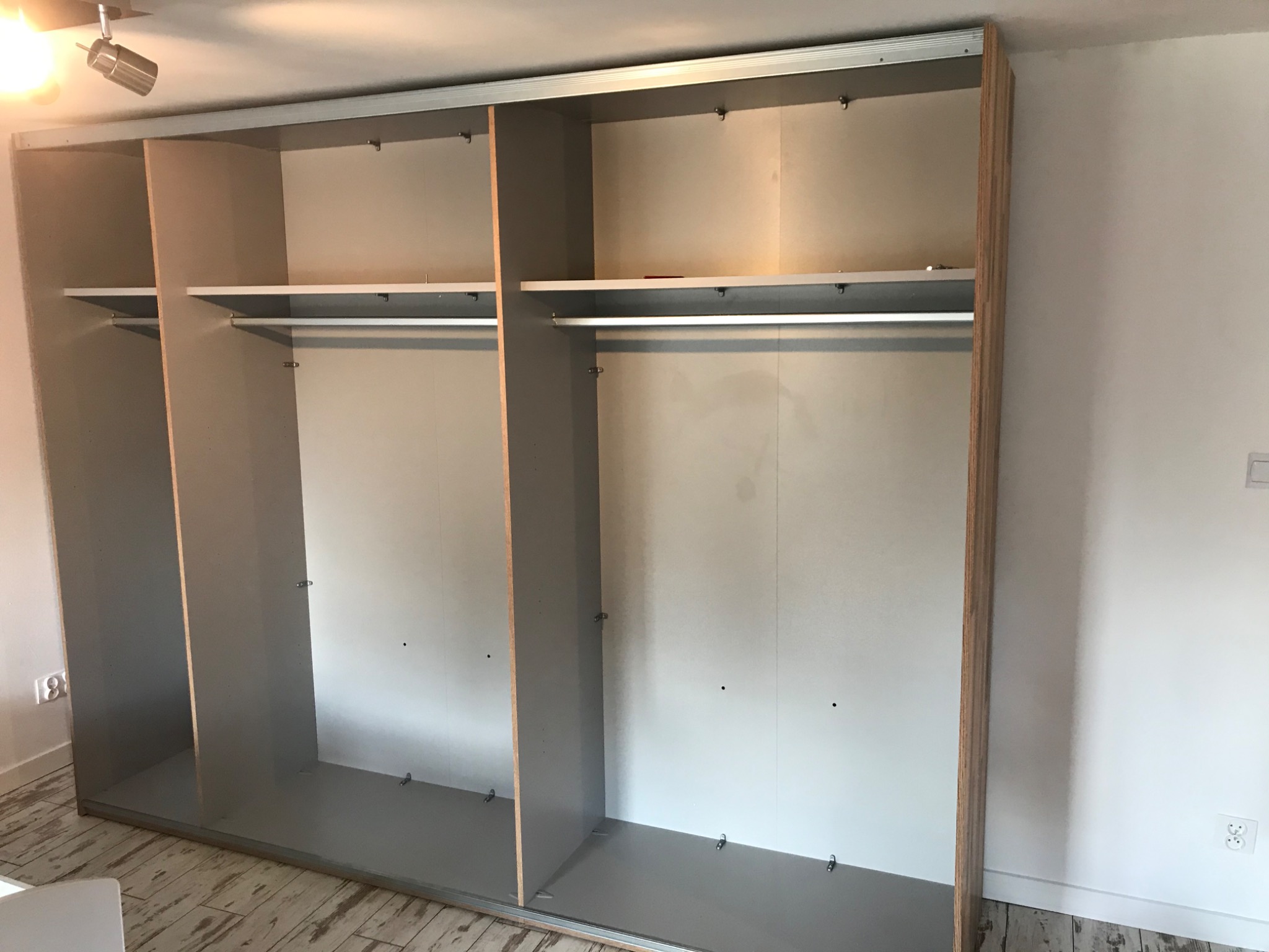 Wnętrze szafy przesuwnej z półkami i drążkami, widoczne mechanizmy mocujące, surowe wykończenie wnętrza, drewnopodobna okleina na krawędziach.