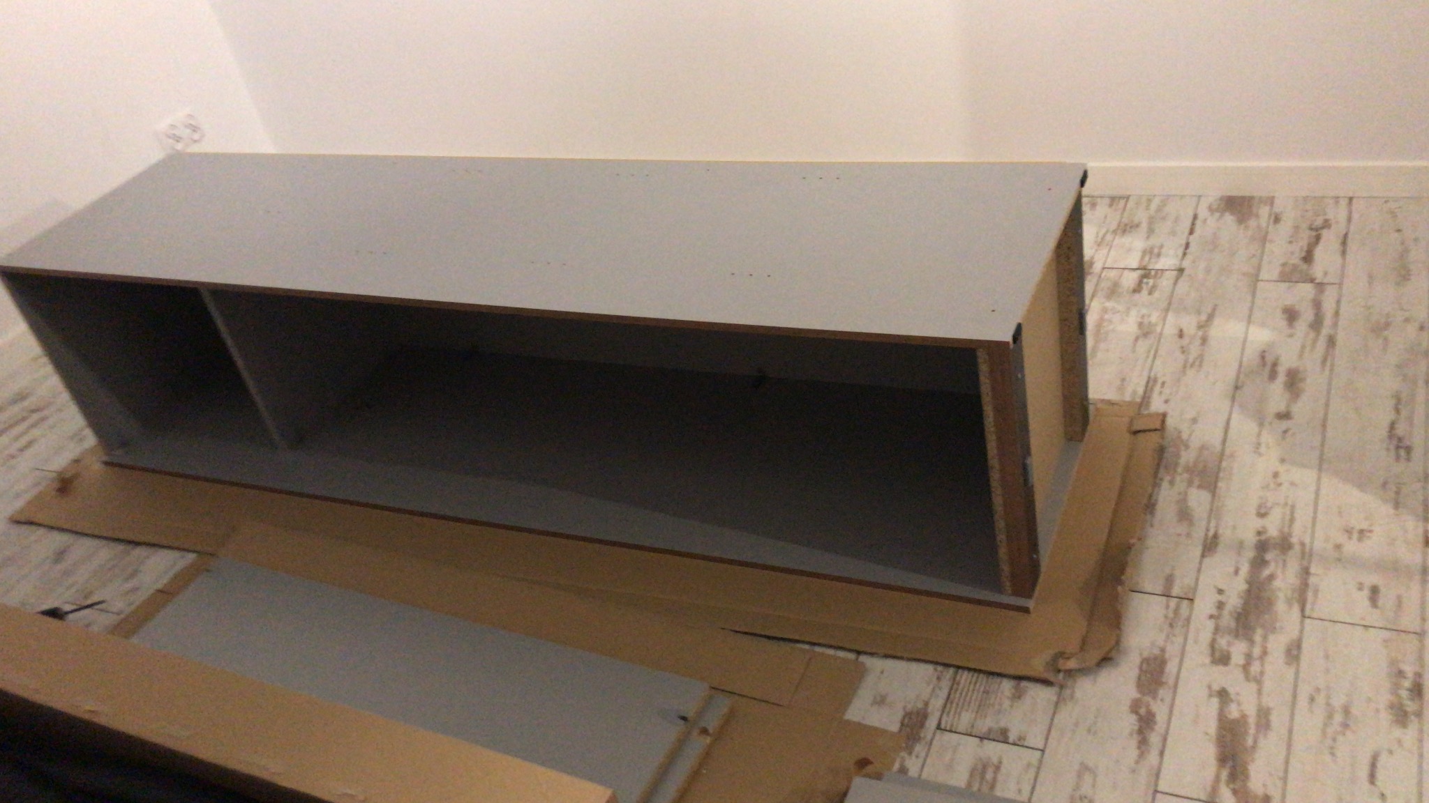 Szara, częściowo złożona szafka z półkami, ustawiona na tekturze na drewnopodobnej podłodze, widoczne otwory na śruby.