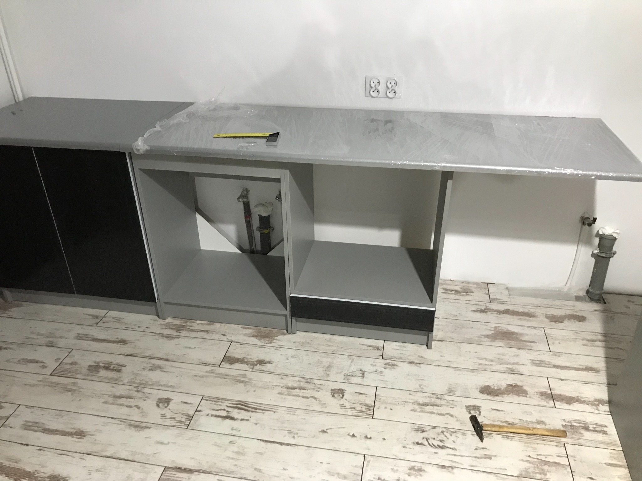Montaż szarej szafki kuchennej z czarnymi frontami, blat zabezpieczony folią, widoczna instalacja hydrauliczna i narzędzie - młotek na podłodze z paneli.