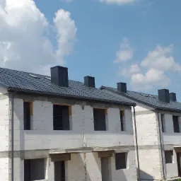 Surowe domy w zabudowie szeregowej z szarym dachem z blachodachówki i czarnymi kominami, w stanie deweloperskim, widoczne rynny.