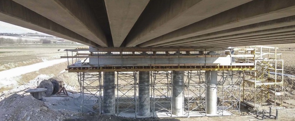 Budowa mostu: widok od dołu na betonowe filary i konstrukcję nośną, z widocznym szalunkiem i rusztowaniem, w tle krajobraz polny.