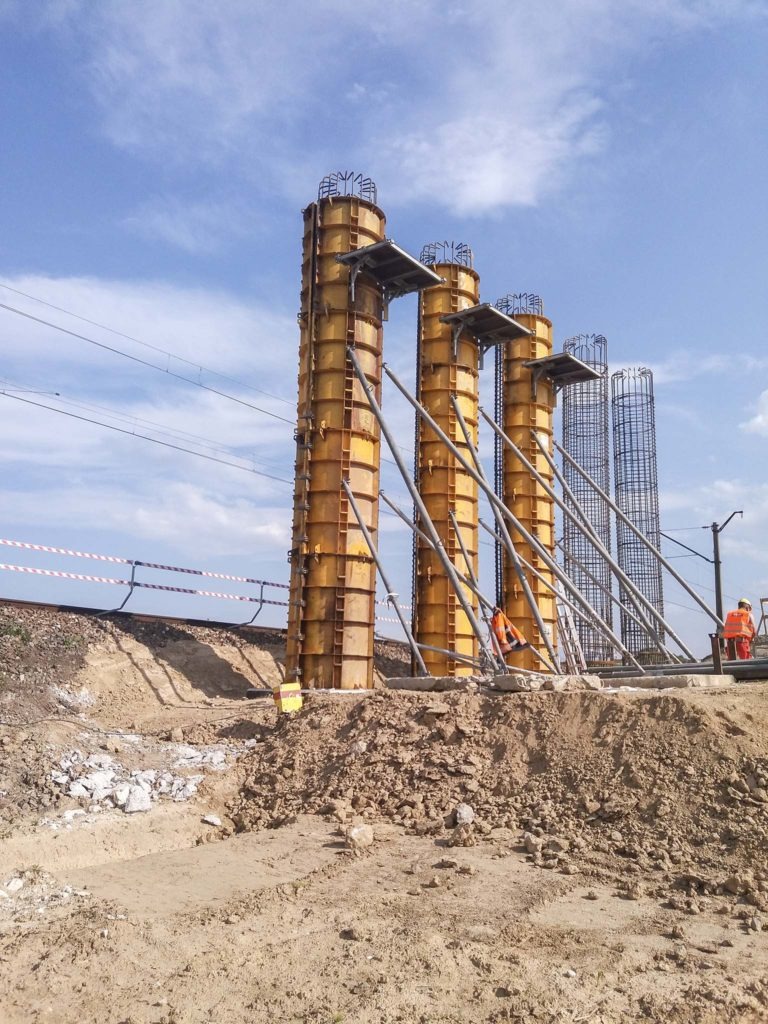 Budowa wiaduktu kolejowego: żółte szalunki kolumn filarów z widocznymi zbrojeniami, dwóch pracowników w pomarańczowych kamizelkach na placu budowy, fragment torów kolejowych w tle.