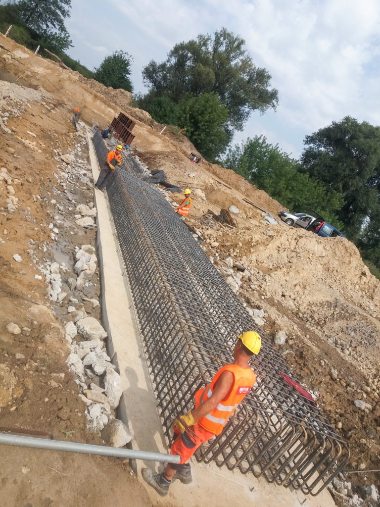 Pracownicy w pomarańczowych kamizelkach układają zbrojenie w wykopie na zboczu, przygotowując podłoże pod konstrukcję drogową.