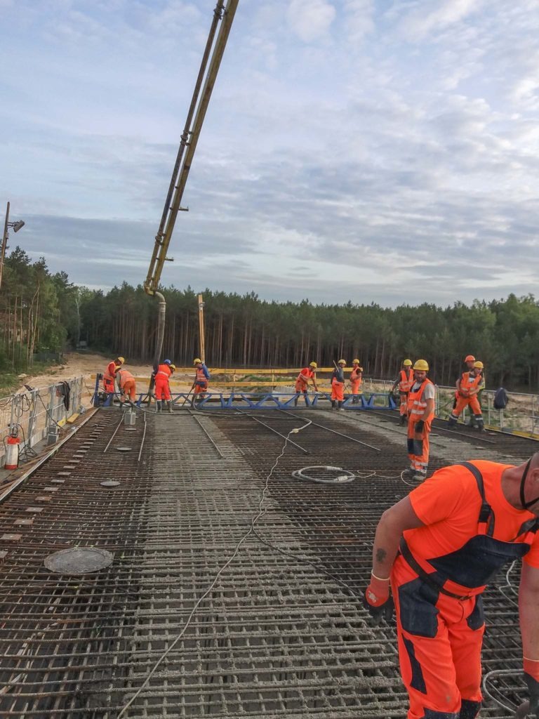 Wylewanie betonu na zbrojenie mostu przez grupę robotników w pomarańczowych kombinezonach, z użyciem pompy do betonu, widok z góry na tle lasu.