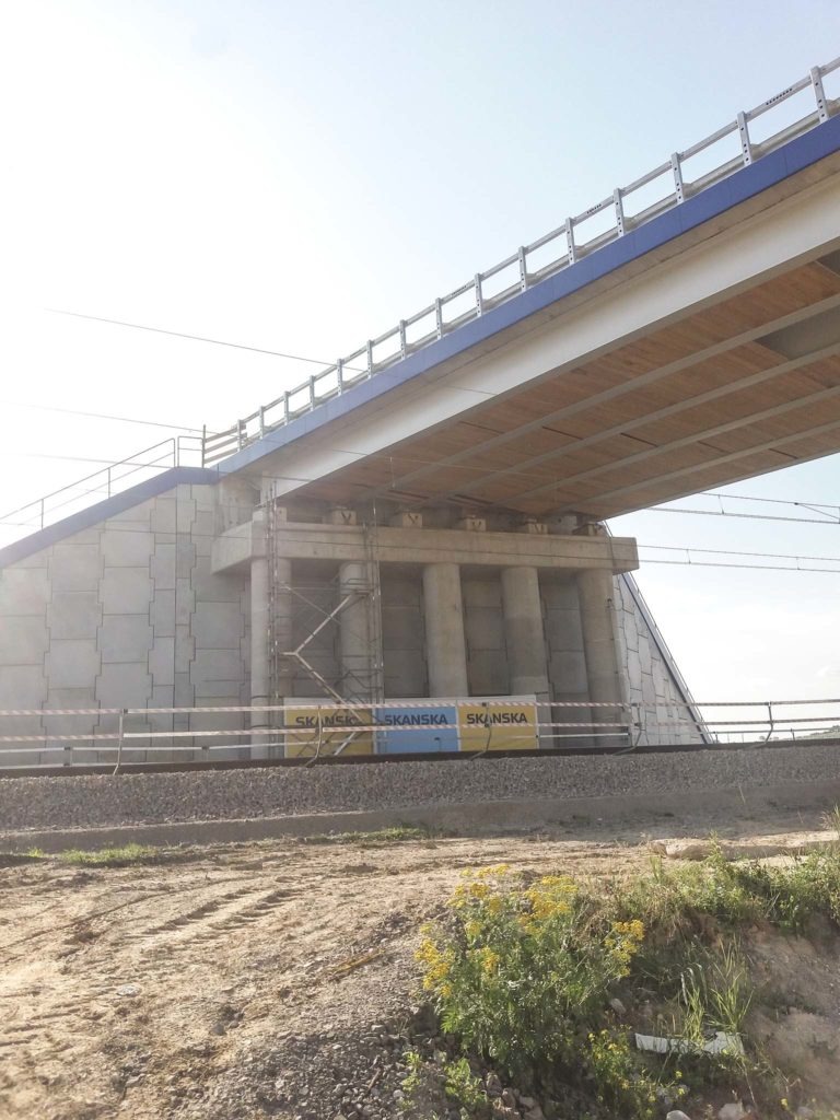 Wznoszący się wiadukt kolejowy z betonowymi podporami, widoczne rusztowania i bariery ochronne, w tle jasne niebo, na pierwszym planie nasyp kolejowy i roślinność.