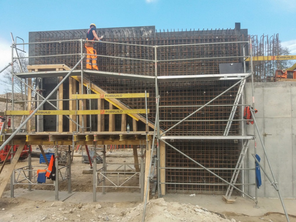 Pracownik w pomarańczowych spodniach na rusztowaniu, zbrojenie pionowej ściany z betonu, widoczne elementy konstrukcyjne i stalowe siatki, teren budowy w słoneczny dzień.