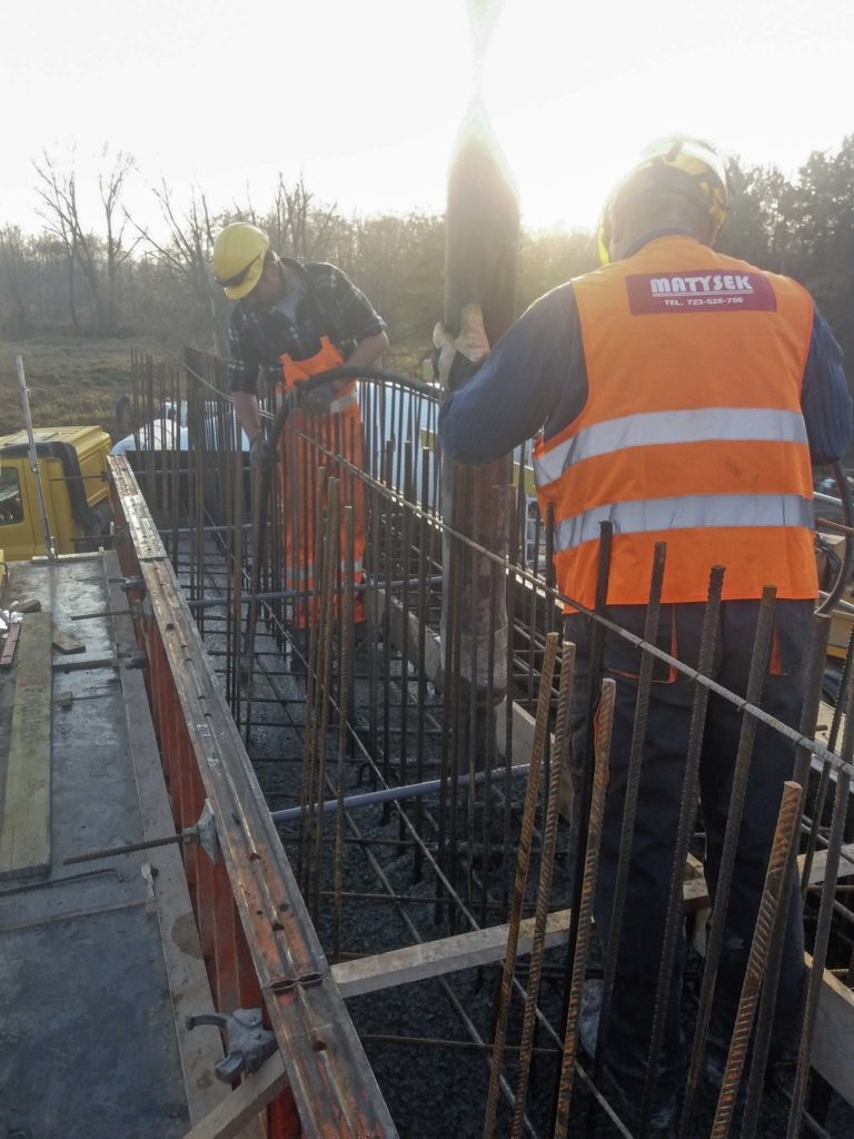 Dwóch pracowników w pomarańczowych kombinezonach i kaskach w trakcie wylewania betonu do szalunku zbrojeniowego na placu budowy w słoneczny dzień.