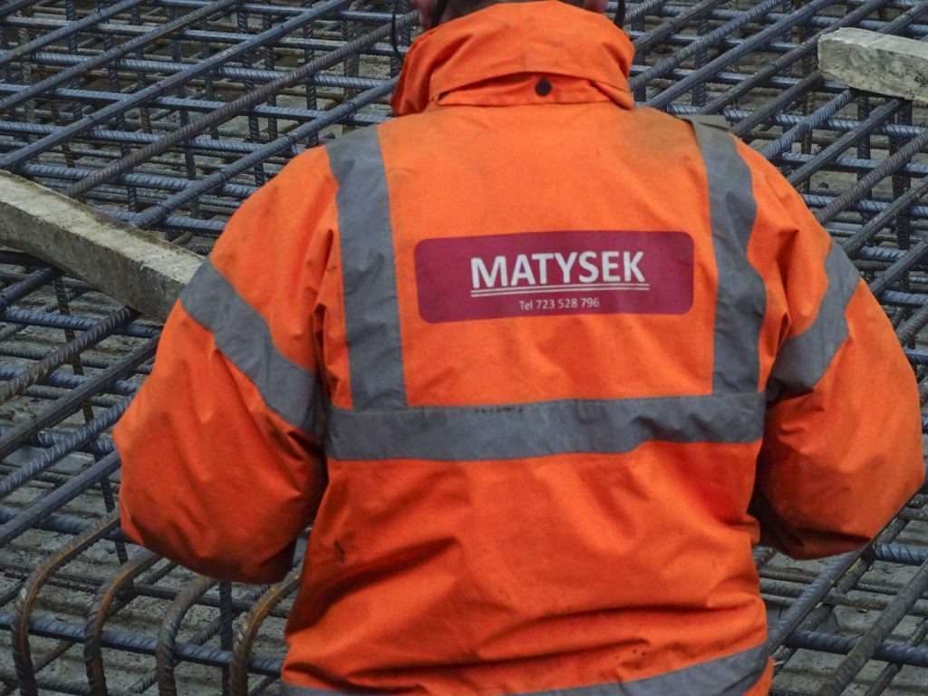 Pracownik w pomarańczowej kurtce z logo firmy MATYSEK na tle zbrojenia fundamentów, widok z tyłu.