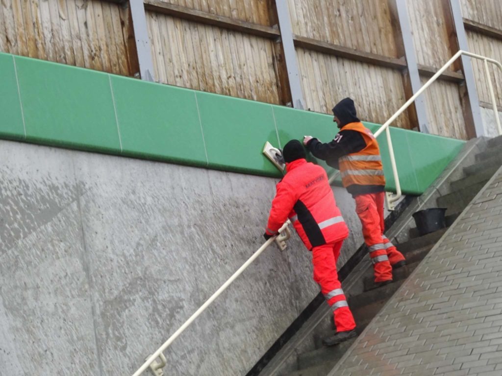 Dwóch pracowników w odblaskowych kombinezonach czyści betonowy mur przy schodach, używając mopa z gąbką i wiadra.
