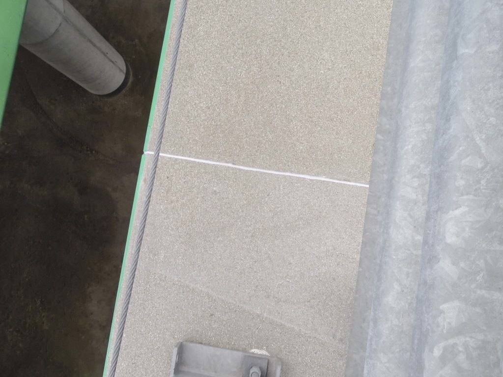 Widok z góry na betonową nawierzchnię z widoczną liną stalową i elementem metalowym, obok fragmentu rury i blachy falistej.