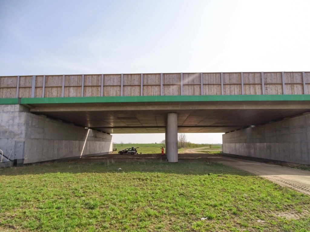 Betonowy wiadukt drogowy z drewnianymi panelami akustycznymi, widoczny z poziomu gruntu, z samochodem i osobą w odblaskowej kamizelce w tle, sugerujące prace budowlane lub inspekcyjne.