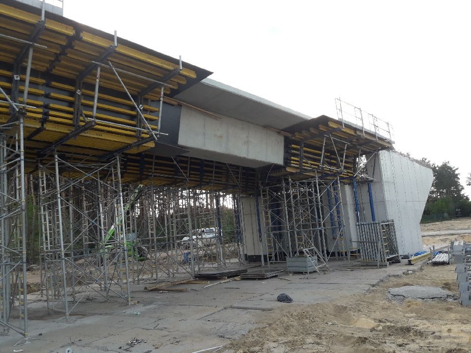 Budowa wiaduktu w Kielcach: widok na konstrukcję wsporczą z rusztowań i żółtych belek pod betonowy element, w tle zieleń drzew.