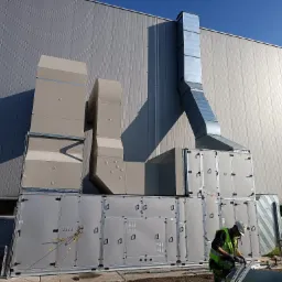 Montaż modułowej instalacji wentylacyjnej z rurami na zewnątrz budynku przemysłowego, pracownik w kasku i kamizelce odblaskowej przycina element konstrukcyjny.