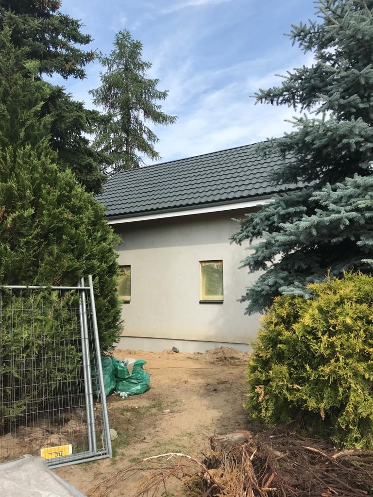 Surowy stan domu szkieletowego z szarą blachodachówką, widoczne zabezpieczone okna, teren wokół domu częściowo uporządkowany, zieleń iglasta w tle.