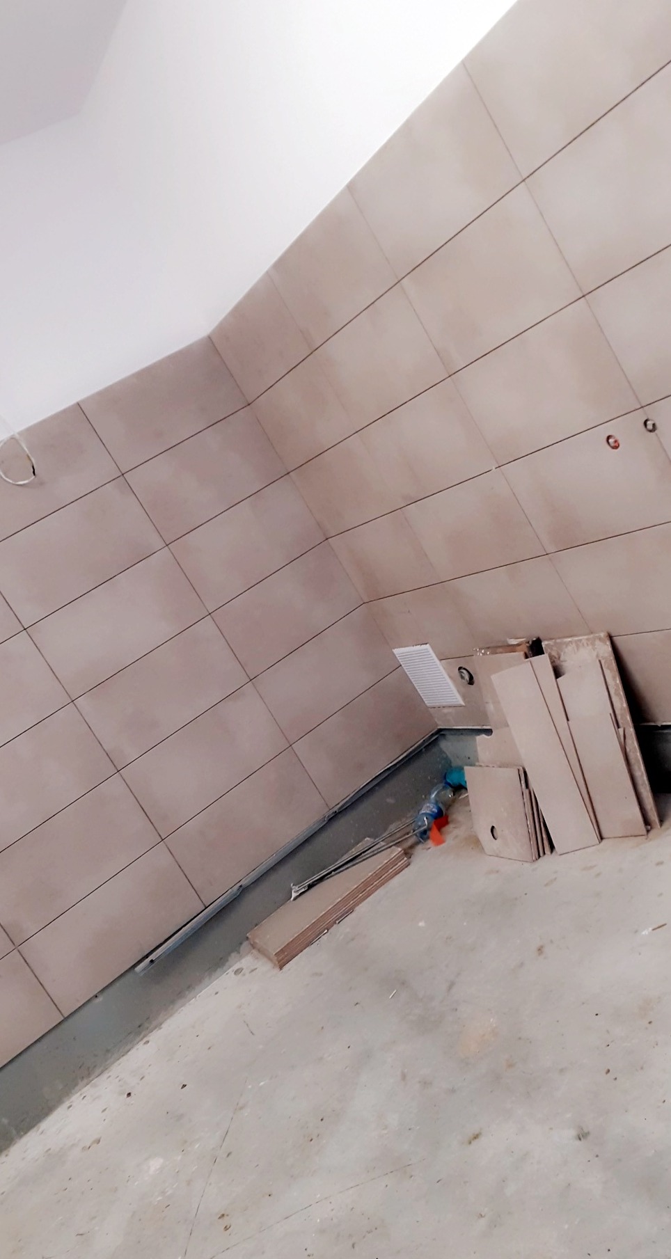 Ściana łazienki wyłożona jasnymi, kwadratowymi płytkami ceramicznymi, widoczne puszki elektryczne, fragment wentylacji, na podłodze płyty kartonowo-gipsowe i resztki materiałów budowlanych.