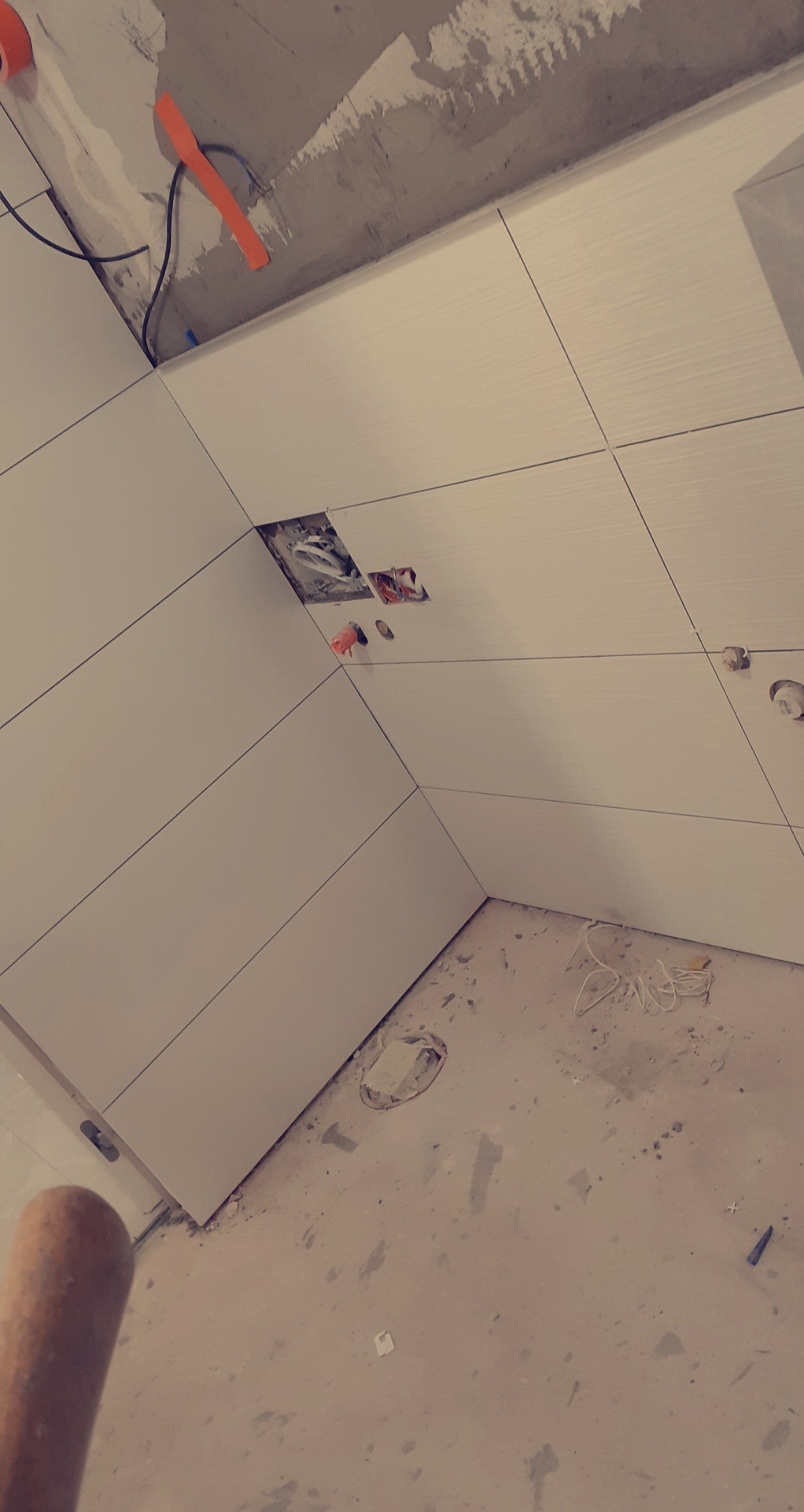 Białe, podłużne płytki ułożone na ścianie, z widocznymi otworami na gniazdka elektryczne i niedokończonymi fugami, surowy betonowy sufit z wystającymi kablami, remont w trakcie.