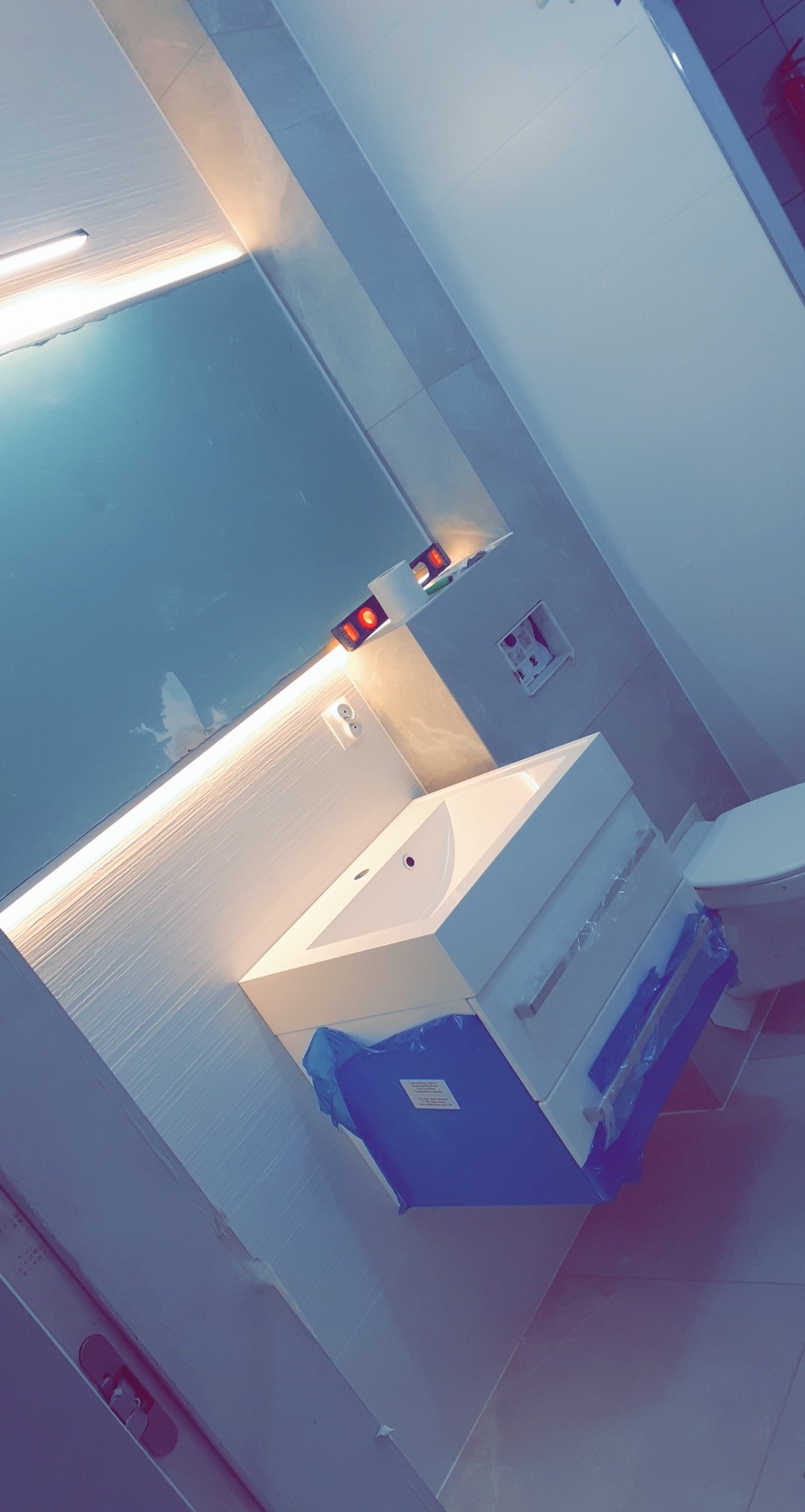 Nowa biała umywalka z szafką, jeszcze zabezpieczona folią ochronną, zamontowana w łazience z jasnymi płytkami, widoczne oświetlenie LED nad lustrem.