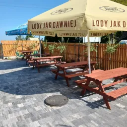 Ustawione rzędem drewniane stoły piknikowe z ławkami na szarym brukowanym placu, osłonięte beżowym parasolem z napisem 'Lody jak dawniej' i logo firmy.