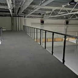 Wnętrze hali produkcyjnej z antresolą zabezpieczoną balustradą szklaną z czarnymi słupkami, widok z góry na szarą posadzkę i białe ściany, oświetlenie przemysłowe.