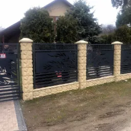 Nowoczesna brama wjazdowa z metalowymi przęsłami o ażurowym wzorze liści, osadzona na beżowych, teksturowanych słupkach z betonowych bloczków, prowadząca na posesję z brukowaną ścieżką.