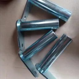 Cztery ocynkowane, metalowe wsporniki z rurką, ułożone ukośnie na płaskiej powierzchni, widoczne ślady po obróbce.