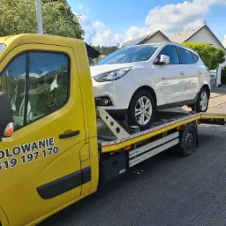 Żółta laweta z białym samochodem osobowym na platformie, widoczne numery kontaktowe na kabinie, tło z domami i zielenią.