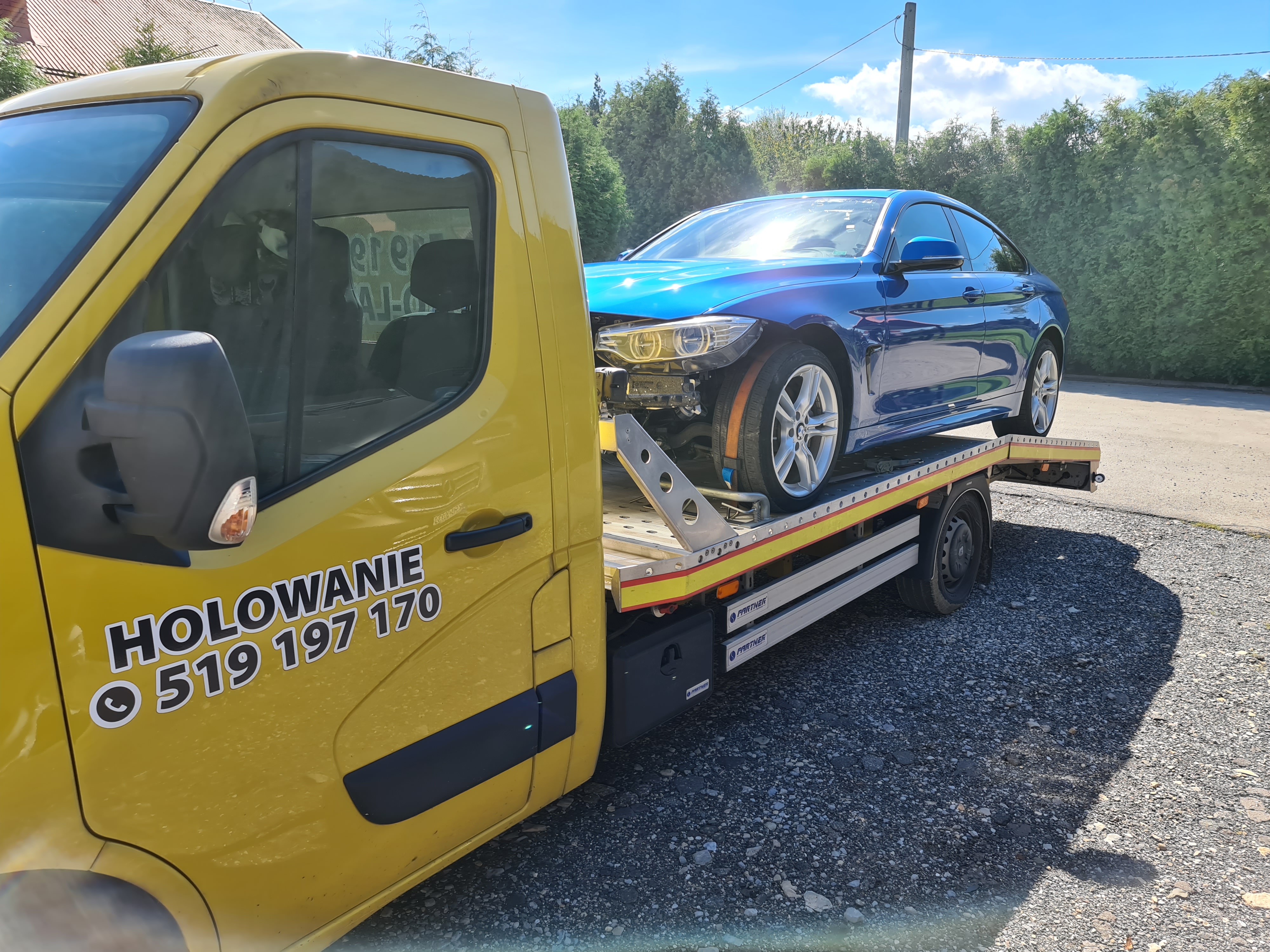 Żółta laweta z uszkodzonym niebieskim samochodem osobowym na platformie, widoczny numer telefonu na kabinie lawety.