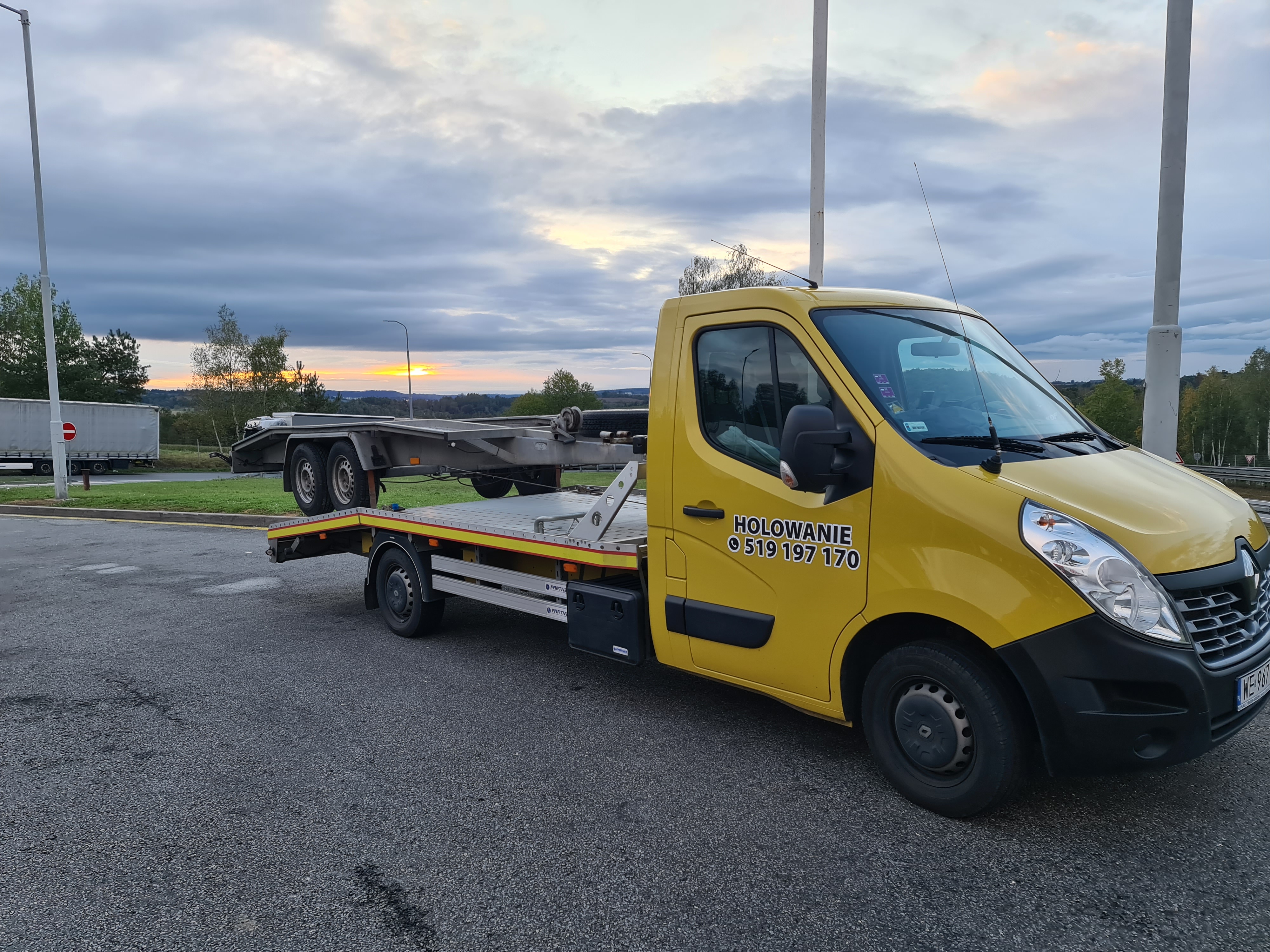 Żółta laweta Renault Master z platformą do przewozu pojazdów, zaparkowana na parkingu z widocznym numerem telefonu na drzwiach, w tle zachodzące słońce.
