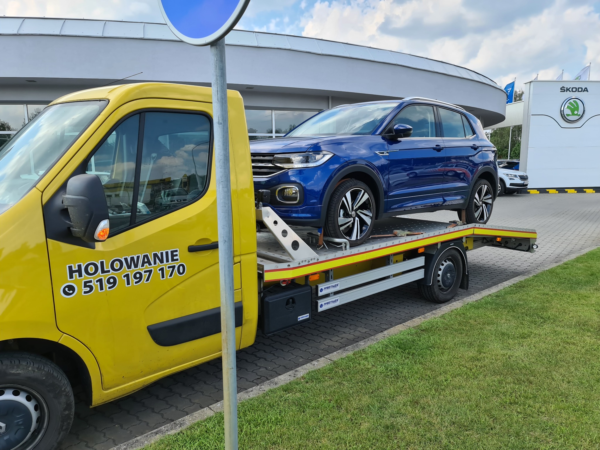 Żółta laweta z niebieskim samochodem osobowym marki Skoda na platformie, na tle salonu samochodowego Skoda.