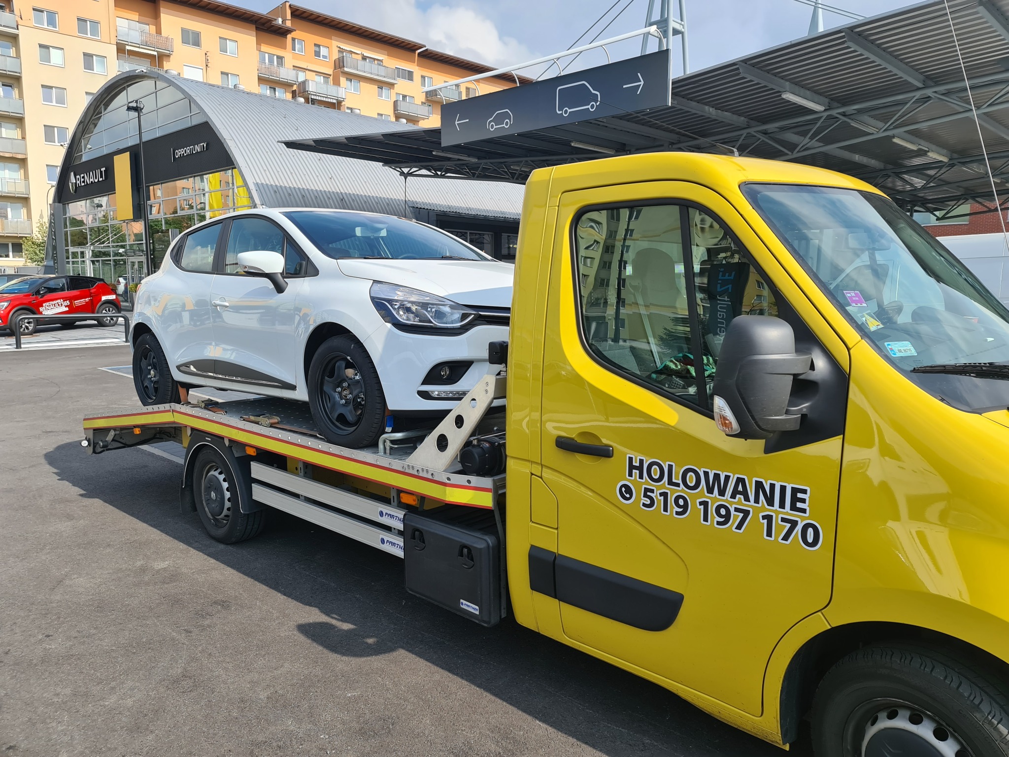 Żółta laweta z białym Renault Clio na platformie, zaparkowana przed salonem Renault z napisem 'Opportunity' na tle budynku mieszkalnego.