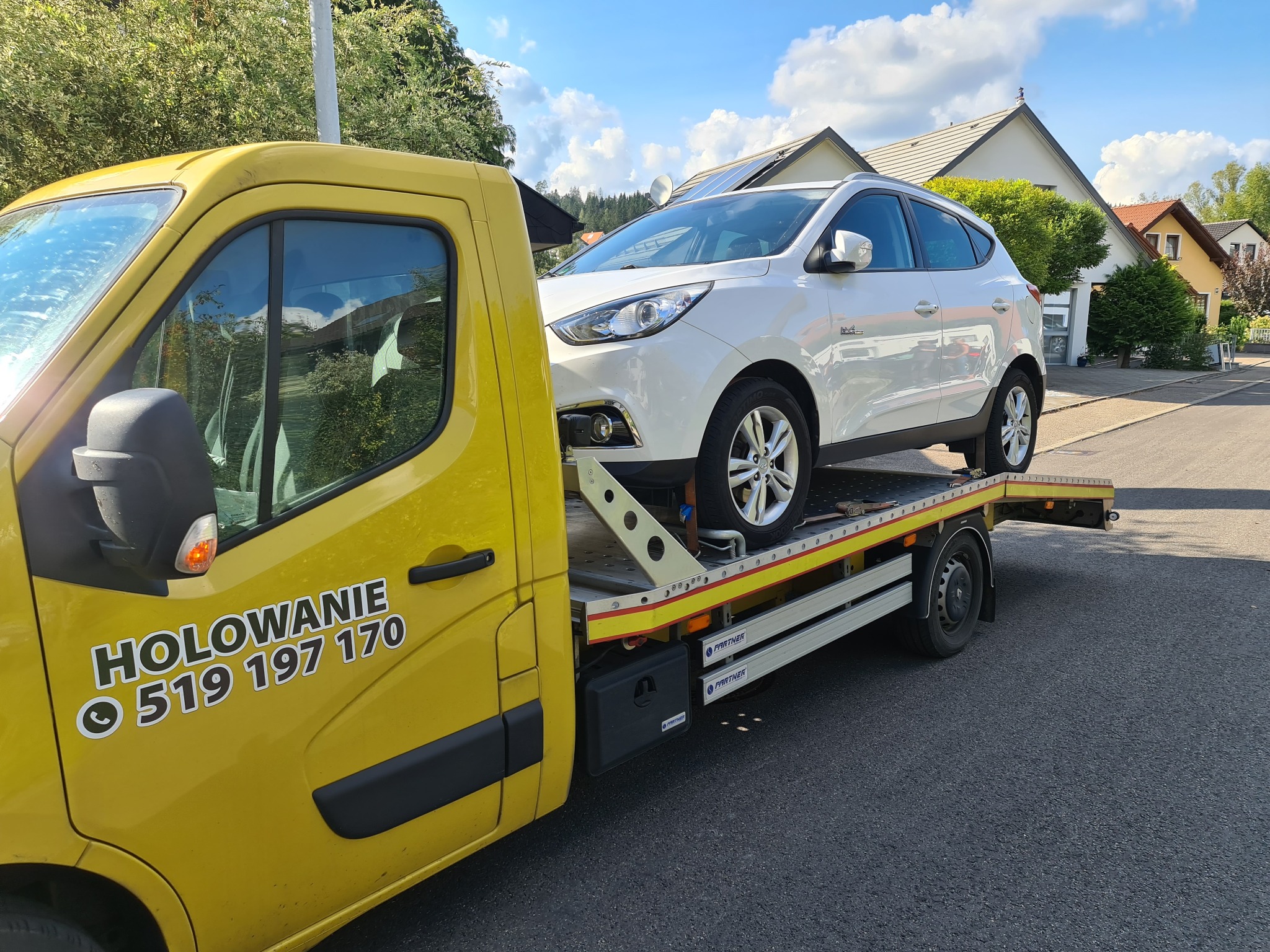 Żółta laweta z białym samochodem osobowym na platformie, numer telefonu 'HOLOWANIE 519 197 170' na drzwiach, domy w tle.