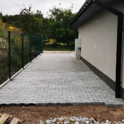 Ułożona kostka brukowa wzdłuż ściany budynku z rynną, widoczna zielona siatka ogrodzeniowa i roślinność w tle, paleta drewniana i kamienie na pierwszym planie.