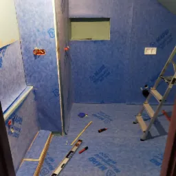 Przygotowanie do hydroizolacji w trakcie remontu łazienki w Ochotnicy Dolnej, widoczne ściany i podłoga pokryte niebieską folią w płynie, poziomica, drabina i inne narzędzia.