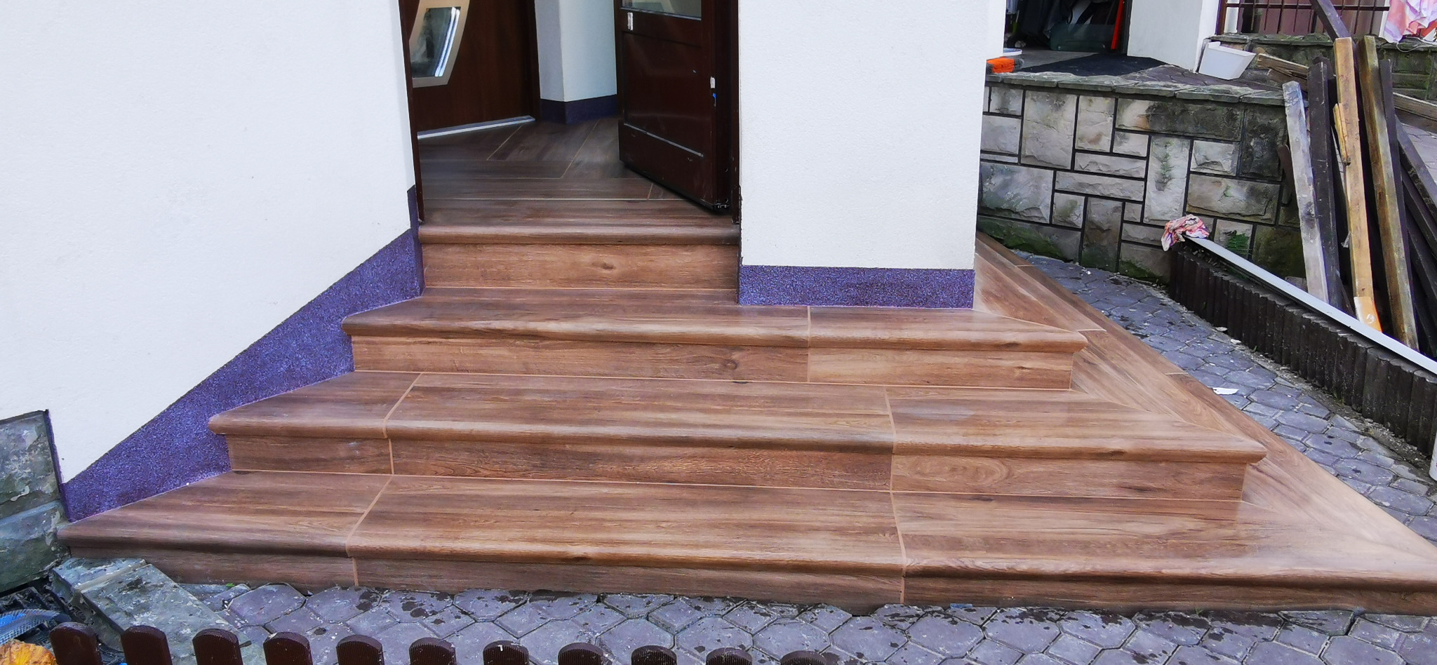 Wykończone schody wejściowe z płytek imitujących drewno, z fioletowym cokolikiem, prowadzące do otwartych drzwi, z widocznym fragmentem brukowanej ścieżki i elementami ogrodu w tle.