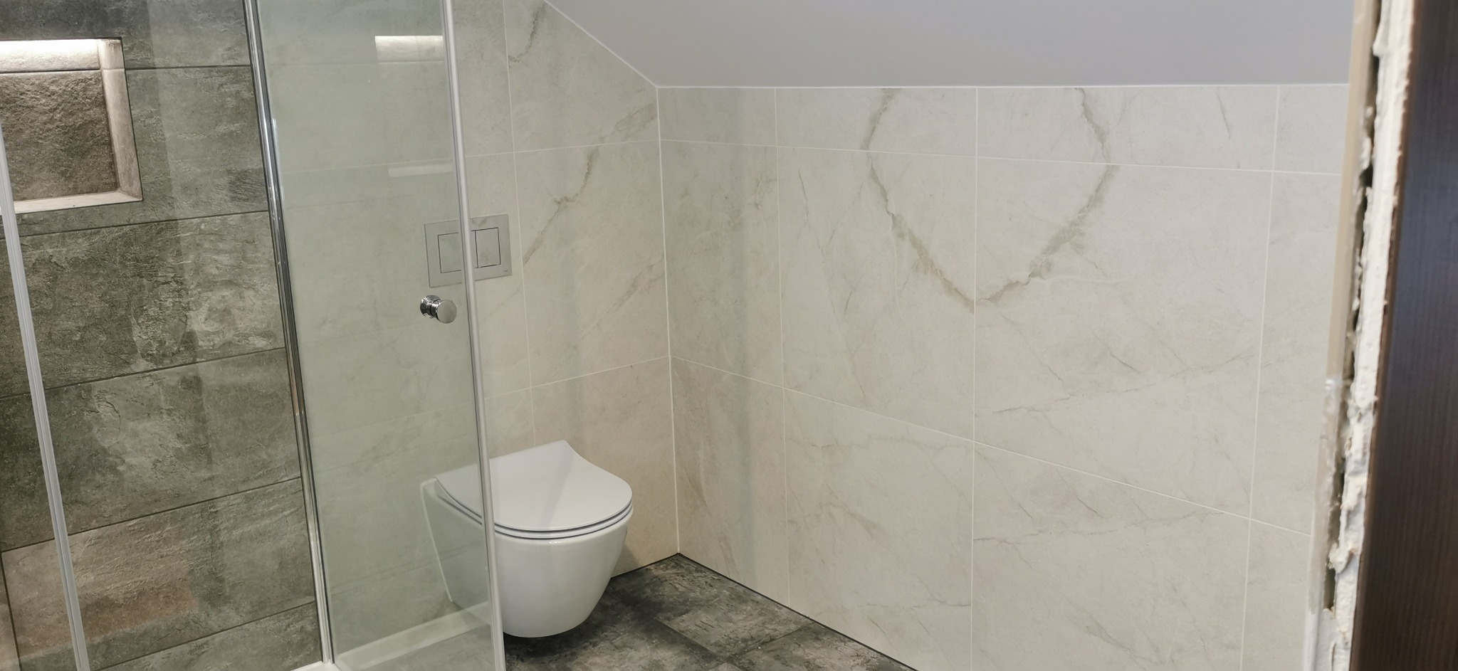 Nowoczesna łazienka z kabiną prysznicową wykończoną szarymi płytkami imitującymi kamień i toaletą podwieszaną, z jasnymi płytkami imitującymi marmur na ścianach, szara podłoga, widoczne prace...