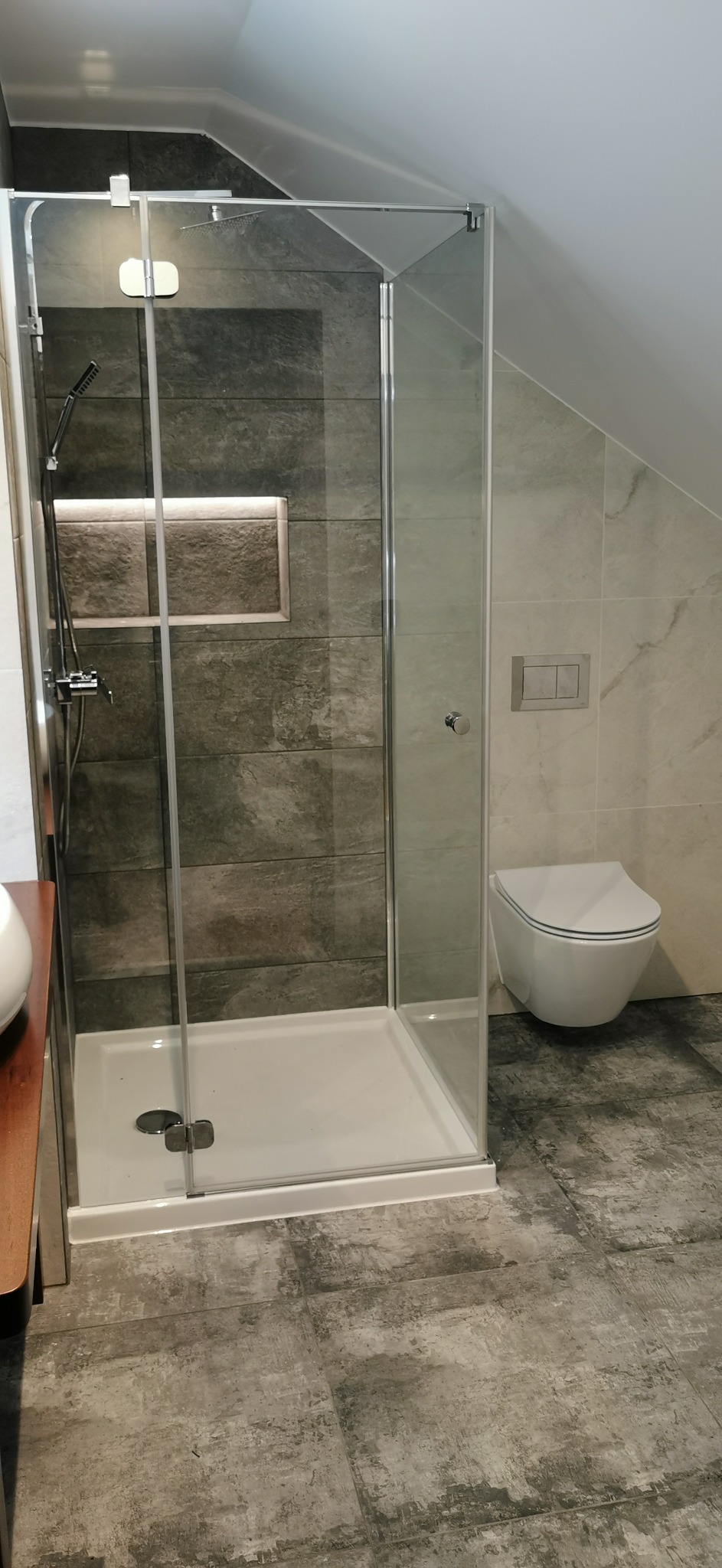 Nowoczesna kabina prysznicowa z chromowanymi okuciami, szara płytka imitująca kamień z podświetlaną wnęką, wisząca toaleta i płytki podłogowe w odcieniach szarości, aranżacja łazienki na poddaszu.