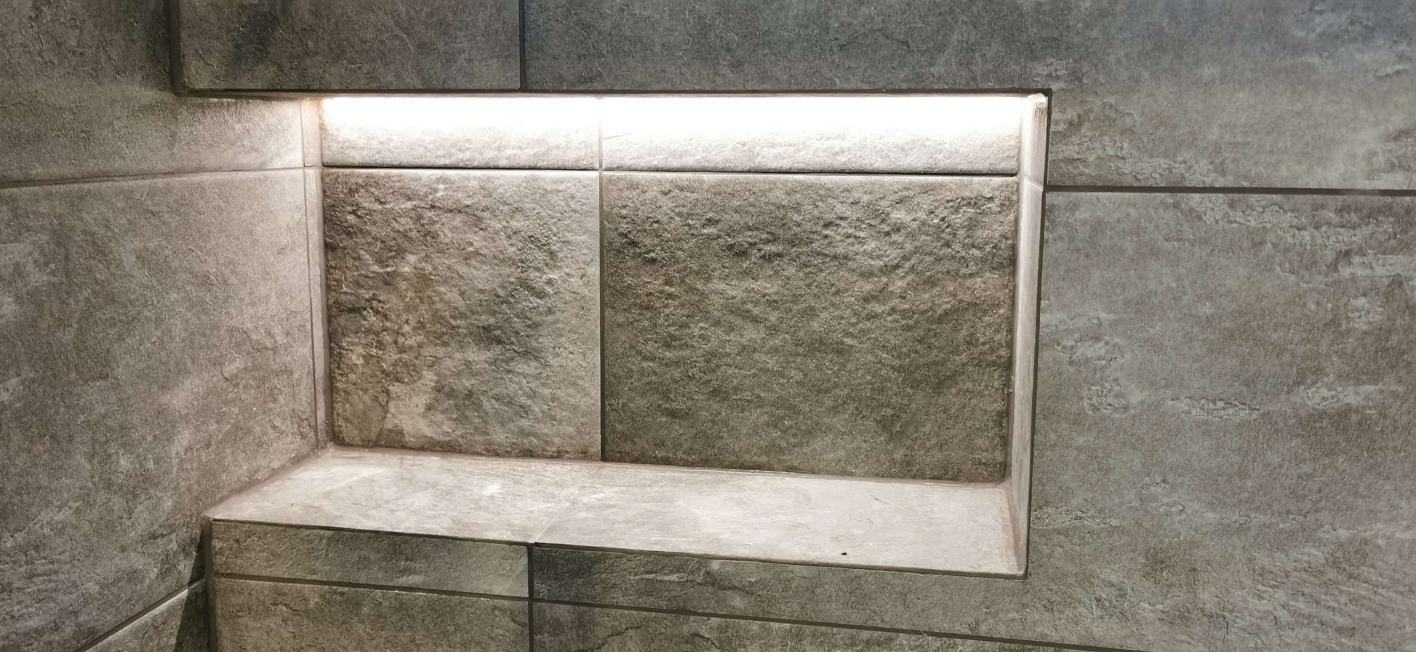 Wnęka prysznicowa wyłożona szarymi płytkami imitującymi kamień, z subtelnym oświetleniem LED w górnej części.