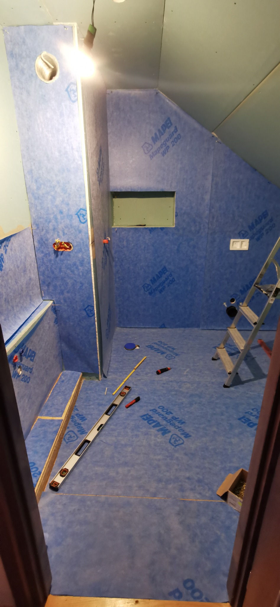 Przygotowanie do hydroizolacji w trakcie remontu łazienki w Ochotnicy Dolnej, widoczne ściany i podłoga pokryte niebieską folią w płynie, poziomica, drabina i inne narzędzia.