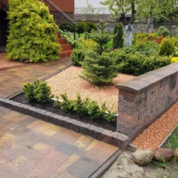 Uporządkowany ogród przydomowy z wybrukowanym chodnikiem, ozdobnymi krzewami, kamienną obwódką i dekoracyjnym żwirem w słoneczny dzień.