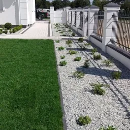 Nowoczesny ogród z równo przystrzyżonym trawnikiem, białymi kamieniami i niskimi roślinami, ogrodzony białym murem z czarnymi, metalowymi przęsłami.