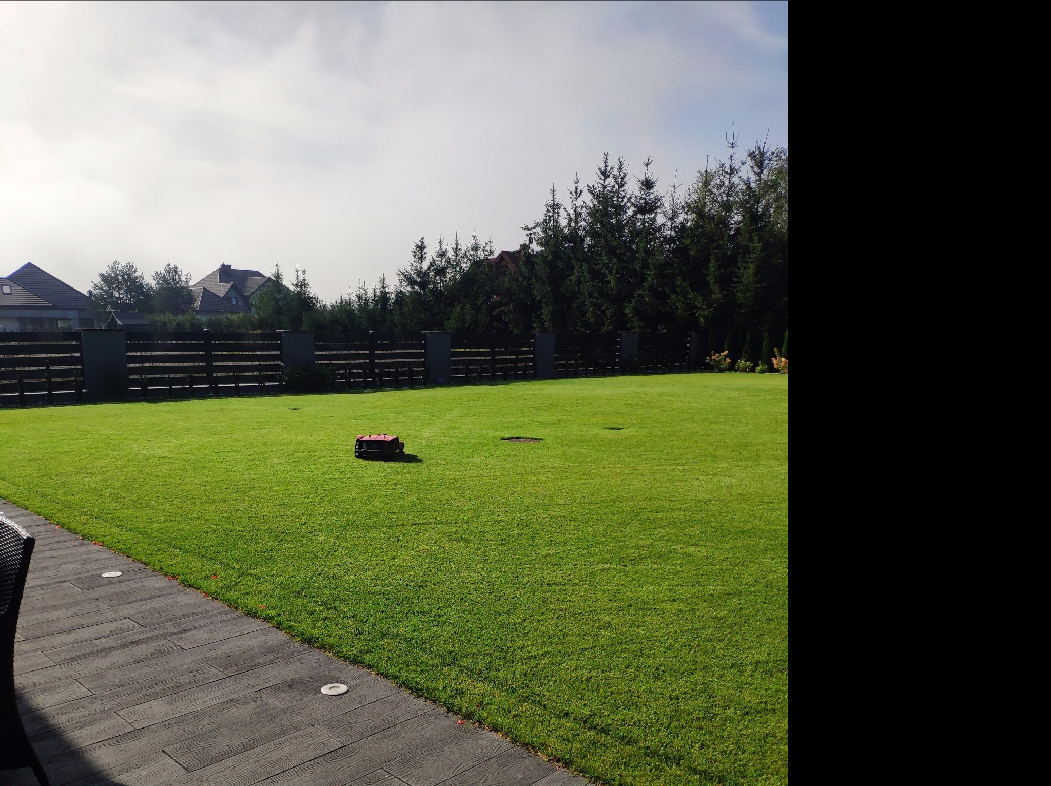 Automatyczna kosiarka do trawy na idealnie przystrzyżonym trawniku przed domem z drewnianym ogrodzeniem i tujami w tle.