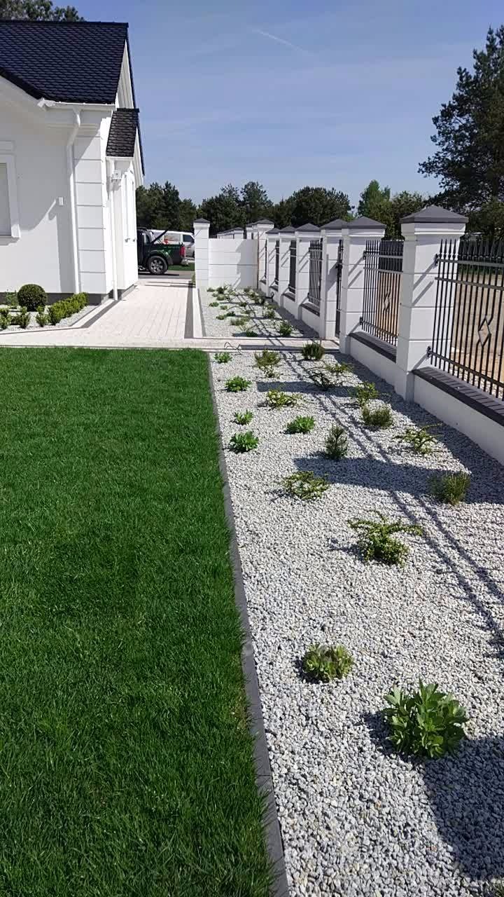 Nowoczesny ogród z równo przystrzyżonym trawnikiem, białymi kamieniami i niskimi roślinami, ogrodzony białym murem z czarnymi, metalowymi przęsłami.